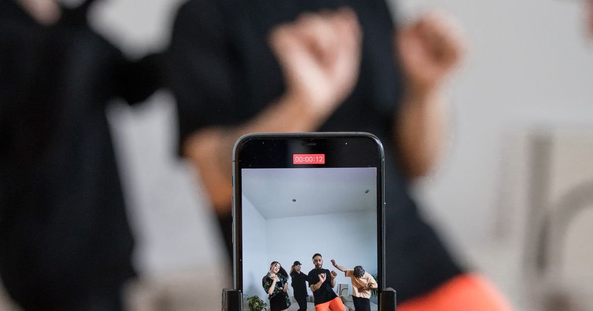 A smartphone on a stand recording a group dancing, with the vertical frame and a recording timer visible on its screen.